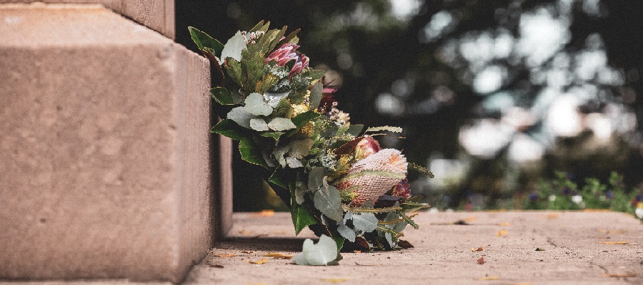 En vakker krans med ulike blomster og grønne blader, plassert ved en steinmur. De jordnære tonene og detaljene i blomstene gir et naturlig og verdig uttrykk, med en uskarp bakgrunn som skaper dybde i bildet. Foto.